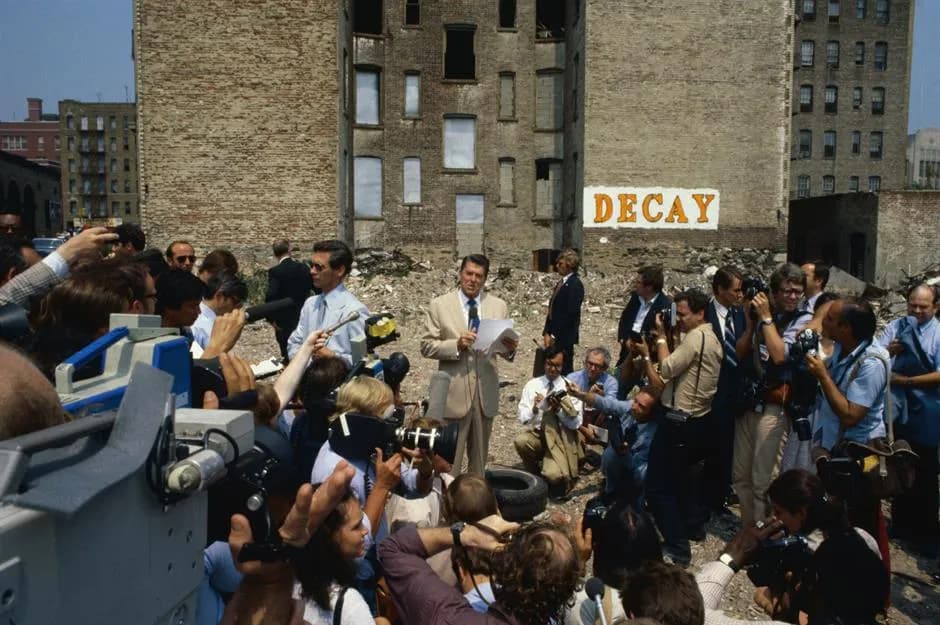 Press crowding a vacant lot beside a sign reading “Decay”