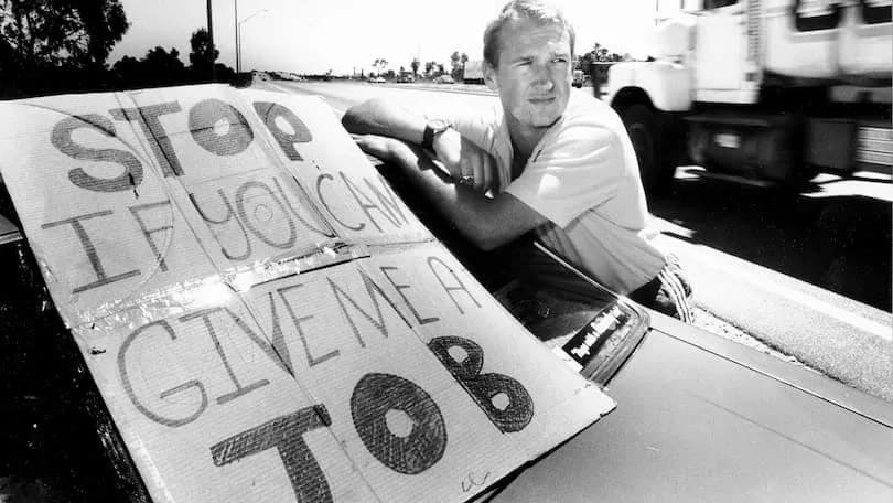 A man leaning on a hand-painted sign reading “Stop if you can give me a job”